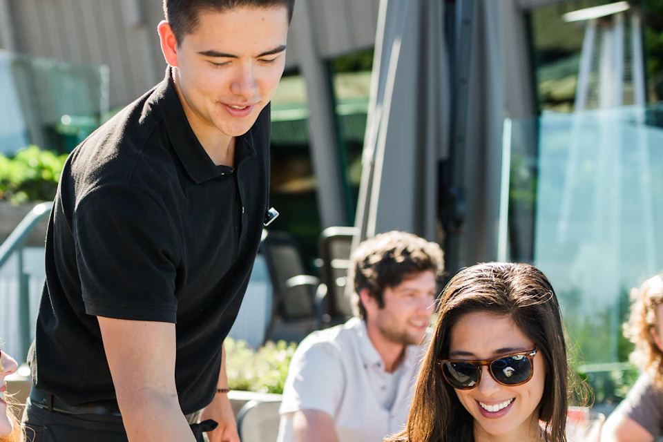 A waiter serves food to smiling customers at an outdoor restaurant; several people are engaged in conversation at nearby tables.