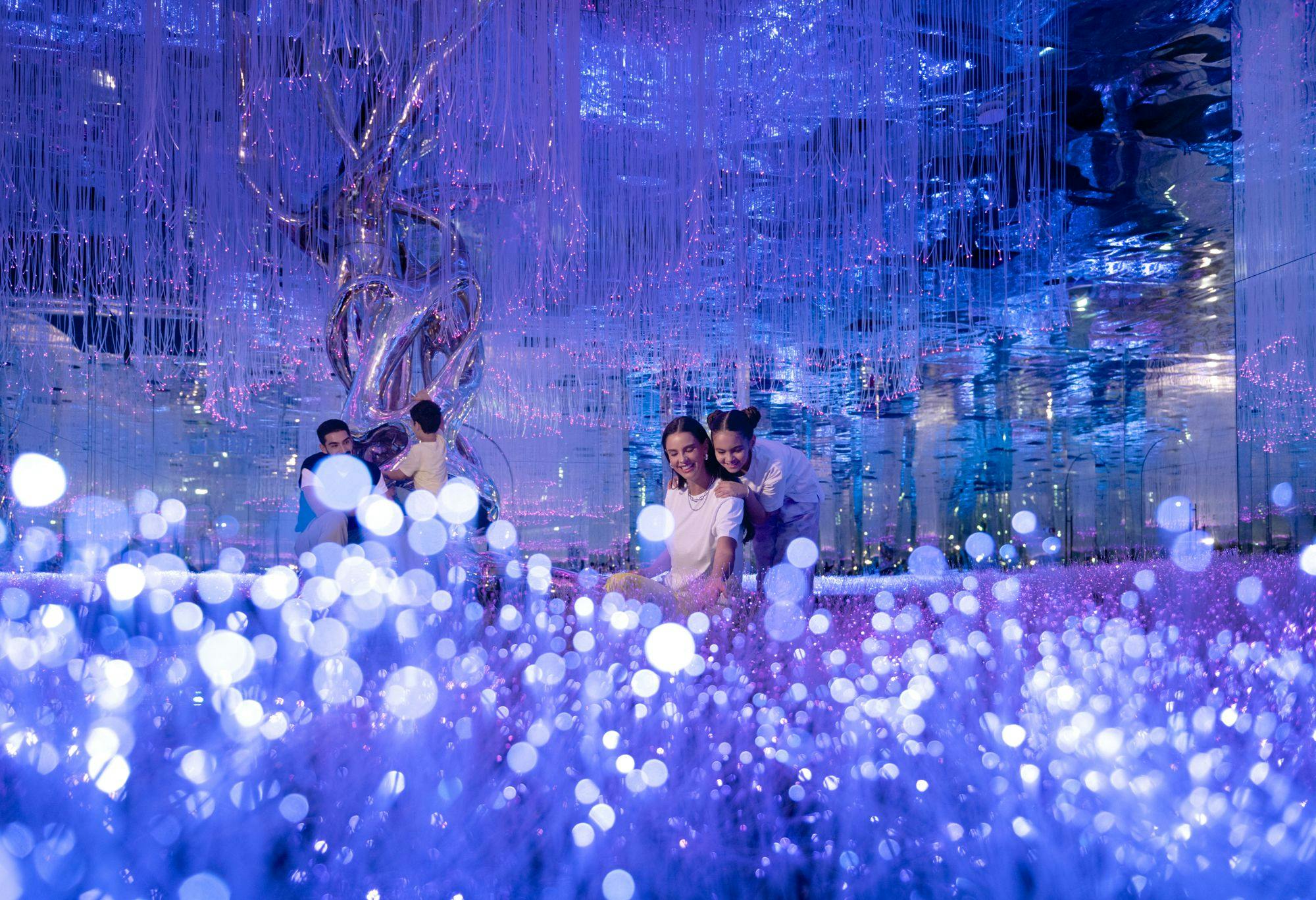 在蓝色灯光的沉浸式艺术装置中，一个女人和孩子在发光的灯光和反光装饰中微笑。