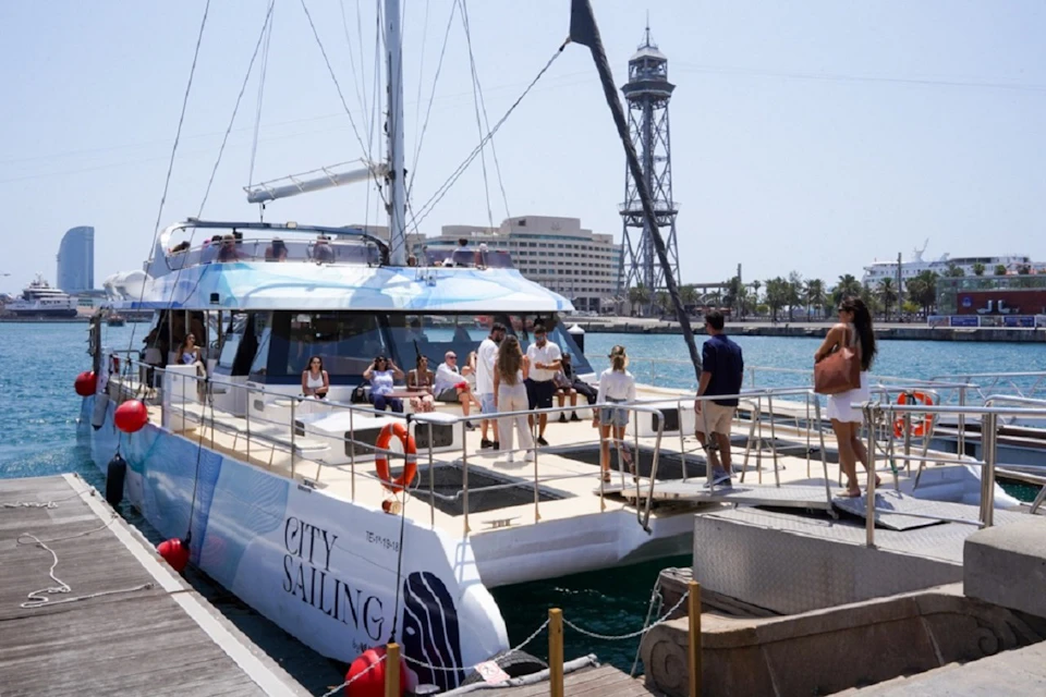 A group of people on a docked catamaran named 'City Sailing,' with a tower and buildings visible in the background.