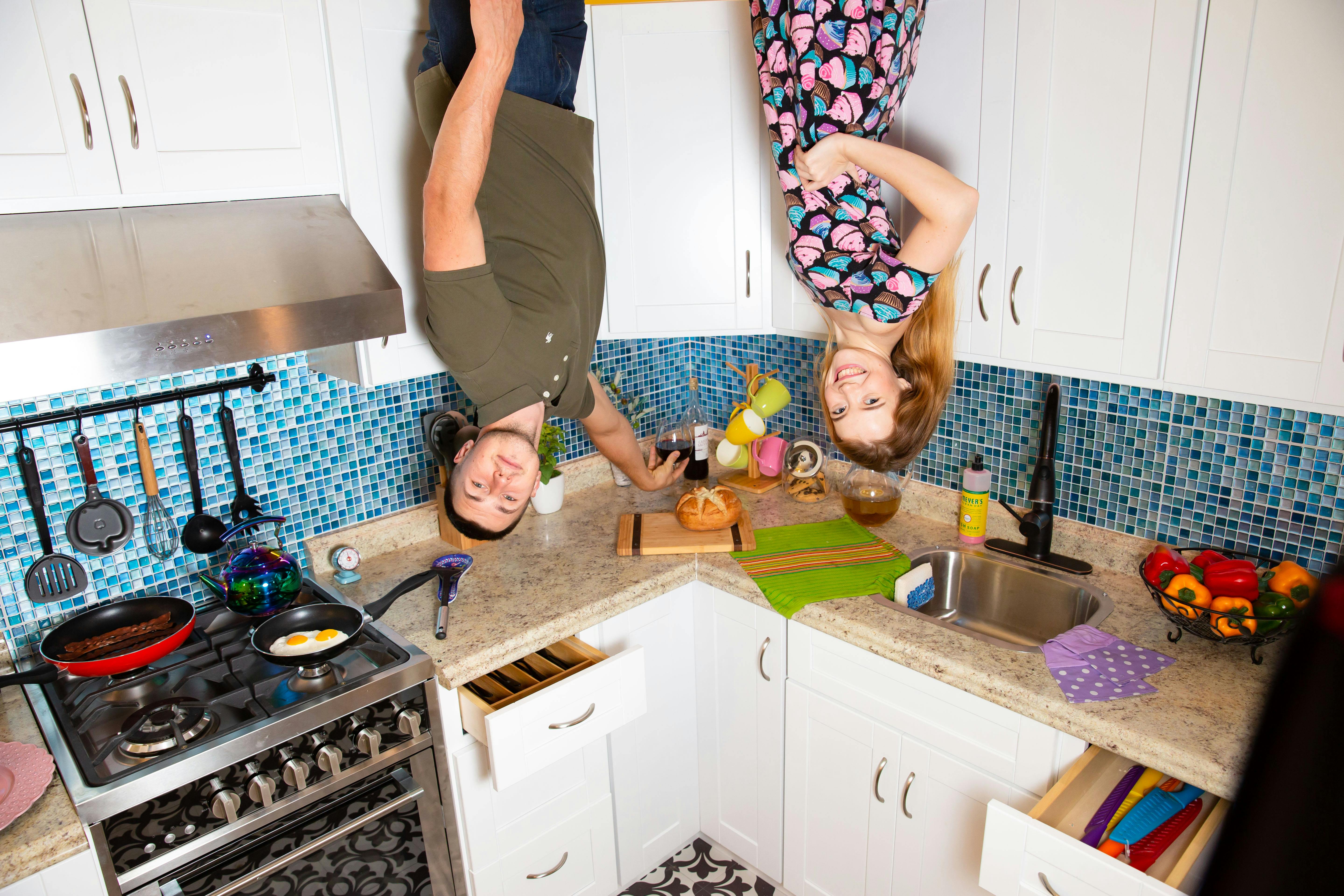 A man and woman hang upside down in a kitchen with blue tiled backsplash, a stove with cooking pans, and a countertop with food.
