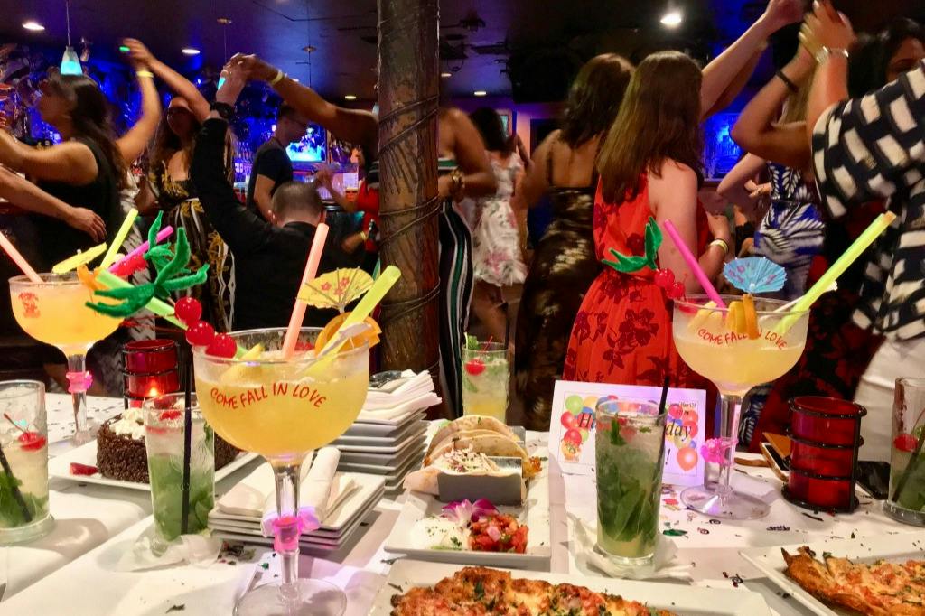 A festive party scene with colorful cocktails, food plates, and people dancing in a lively indoor setting.