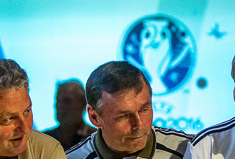 A group of men, some in sports jerseys, gather around an interactive display with a large soccer ball and stadium scene in the background.