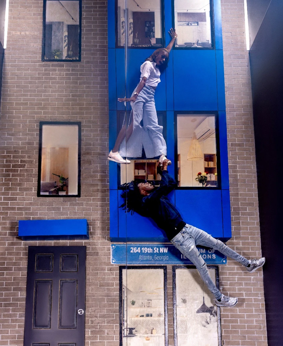 Two people appear to defy gravity, holding onto each other while seeming to float in front of a brick building.