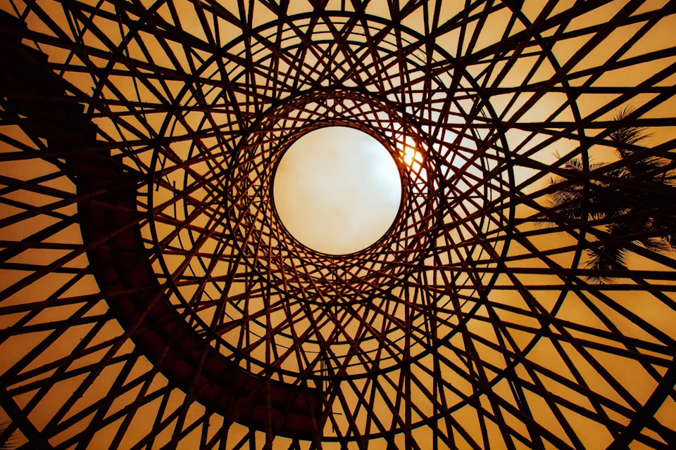 A circular lattice structure viewed from below, with the sun visible through the center against a warm sky.