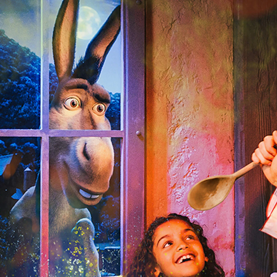 Two girls and a chef stir a glowing, green cauldron with a donkey visible through a window behind them.