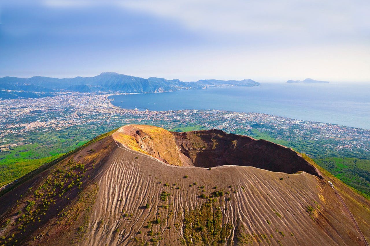 Aerial view of a volcanic crater with ridged, barren slopes, a distant coastal city, and a bay under a clear sky.