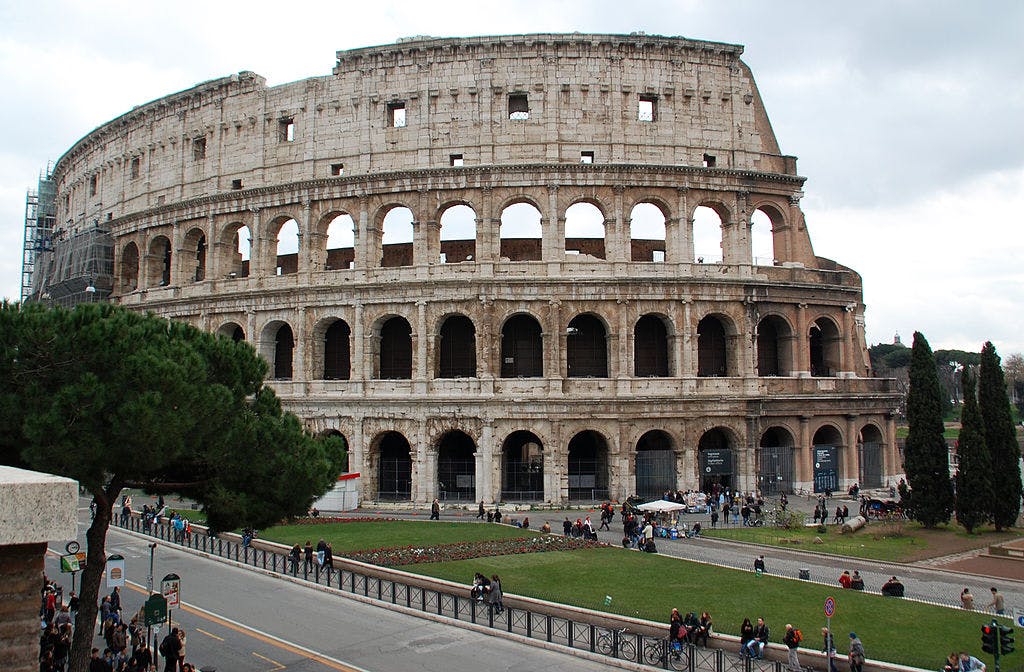 L'immagine mostra il Colosseo romano con molti turisti intorno. Nelle vicinanze sono visibili degli alberi e un'area erbosa.