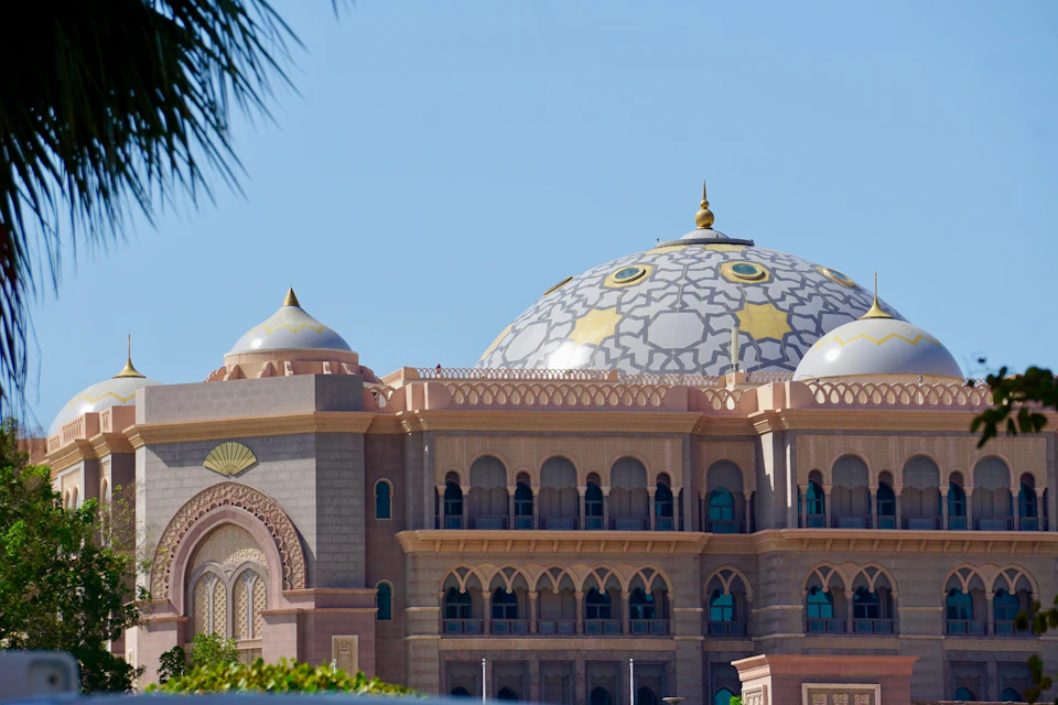 A large ornate building with domes, intricate details, and arched windows, set against a clear blue sky and palm leaves.