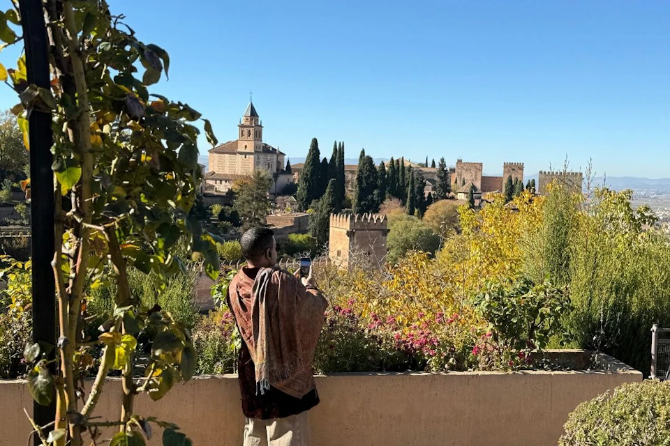A person wearing a shawl takes a photo of a scenic landscape with historic buildings and trees under a clear blue sky.
