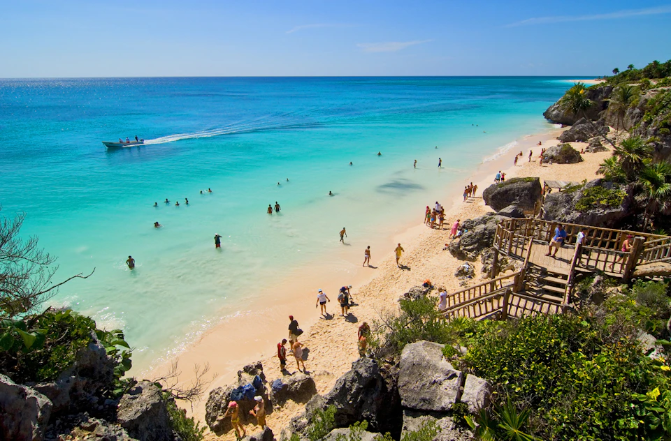 People enjoying a sunny day on a sandy beach with clear turquoise water, some swimming, others strolling or sunbathing, rocky steps nearby.