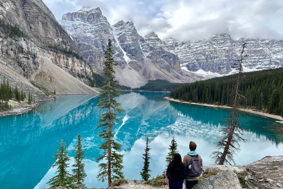 Two people stand overlooking a turquoise lake surrounded by tall pine trees, with snow-capped mountains in the background.