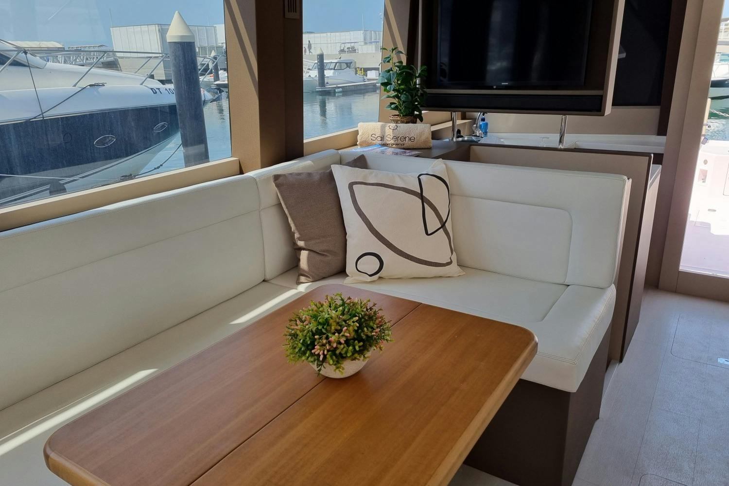 A cozy yacht interior with white seating, decorative pillows, a wooden table, and a potted plant. A marina is visible outside.