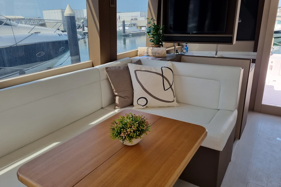 A cozy yacht interior with white seating, decorative pillows, a wooden table, and a potted plant. A marina is visible outside.