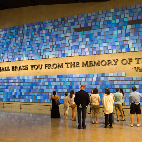 Menschen stehen vor einer Wand aus blauen Kacheln mit der Aufschrift "KEIN TAG WIRD DICH AUS DEM GEDÄCHTNIS DER ZEIT LÖSCHEN - Virgil".