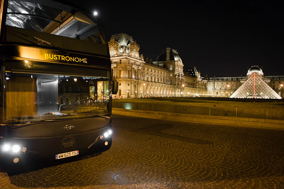 A double-decker bus labelled 'Bustronome' at night near an illuminated historic building with intricate architecture.