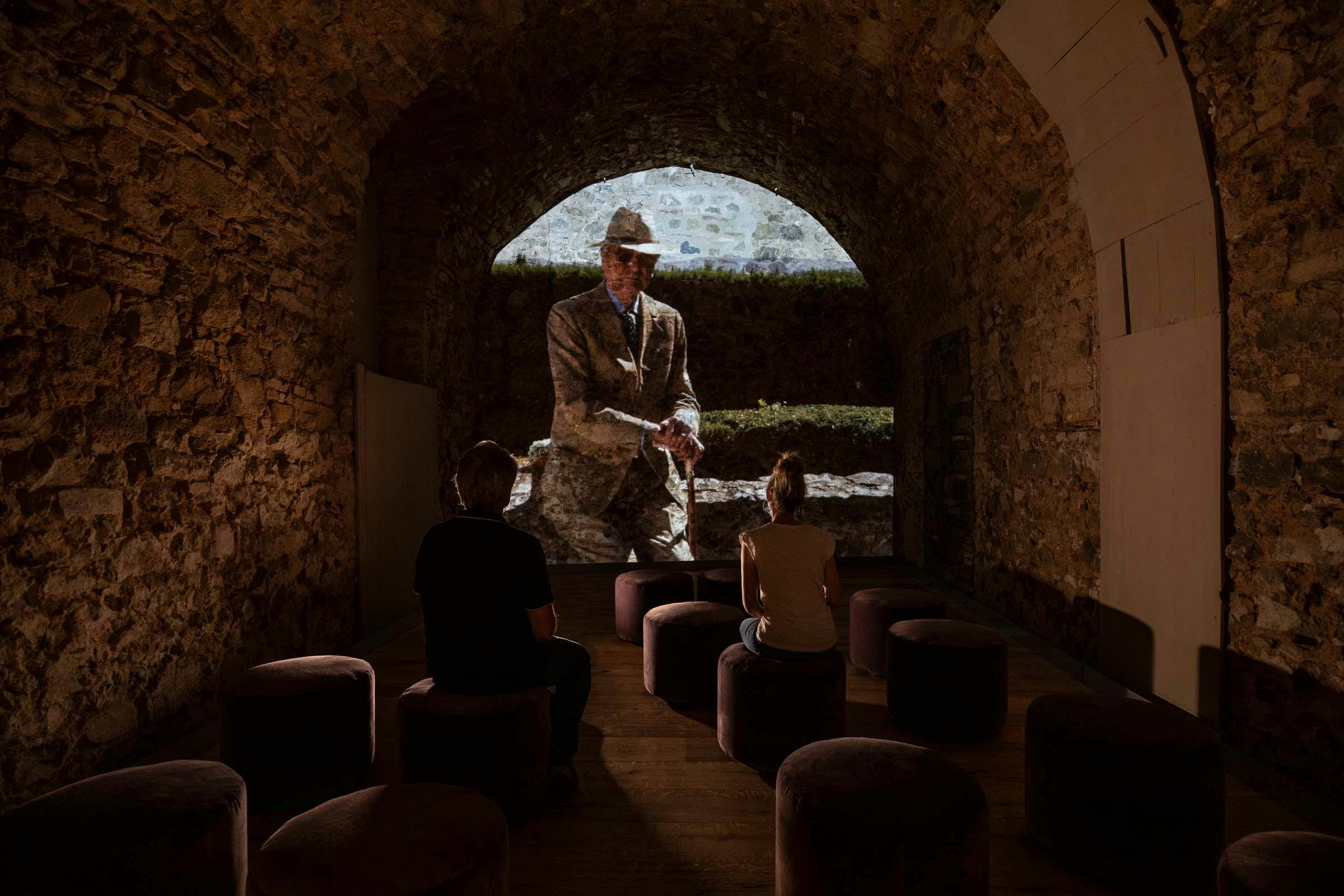 People sit on stools watching a historical projection in a dimly lit, stone-walled room.
