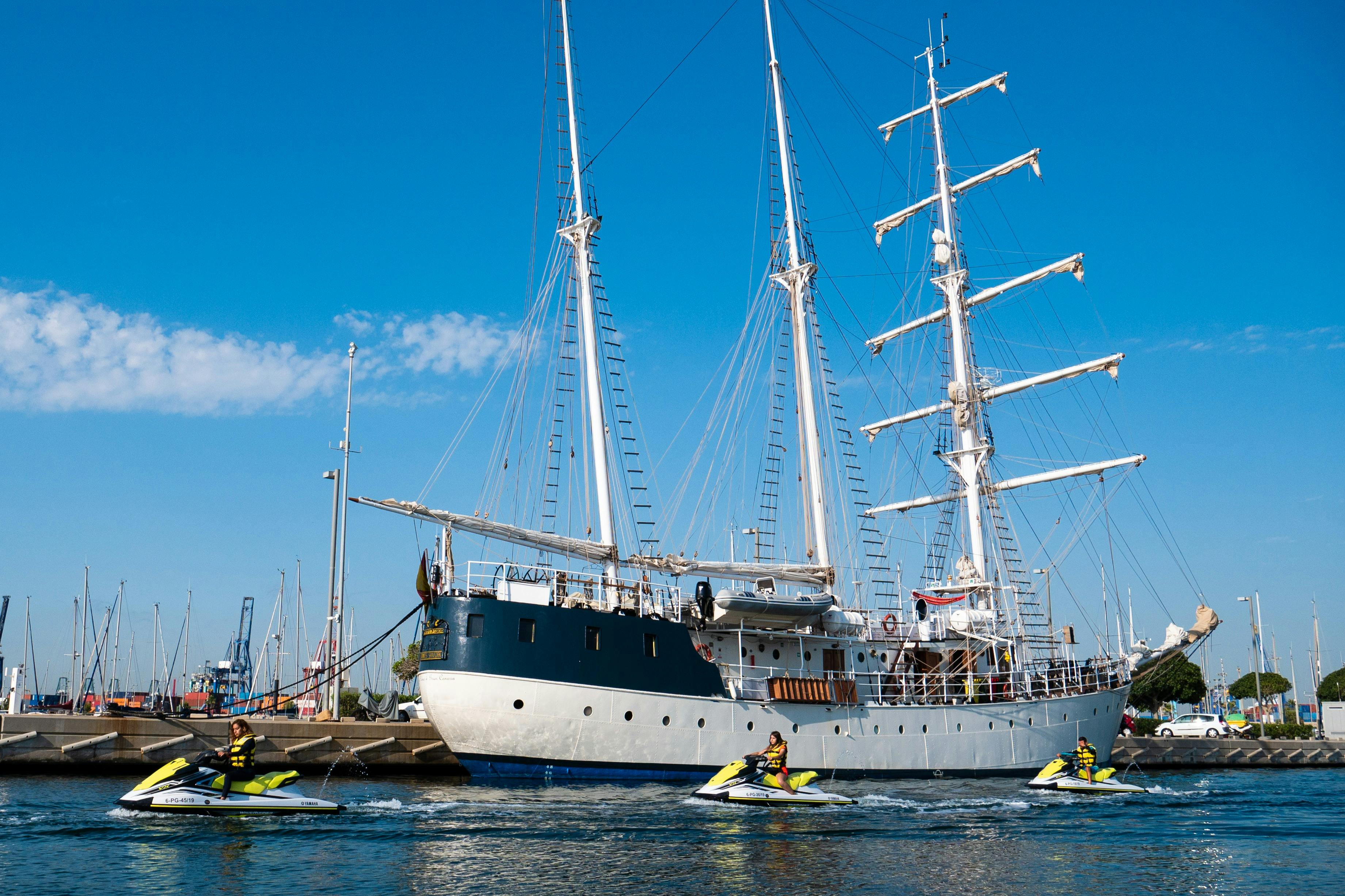 Za slunečného dne zakotvily vedle sebe dvě velké plachetnice. Lidé jezdící na vodních skútrech ve vodě v blízkosti lodí.