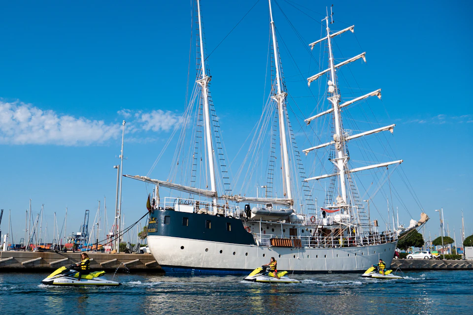 Two large sailboats docked side by side on a sunny day. People riding jet skis in the water near the boats.