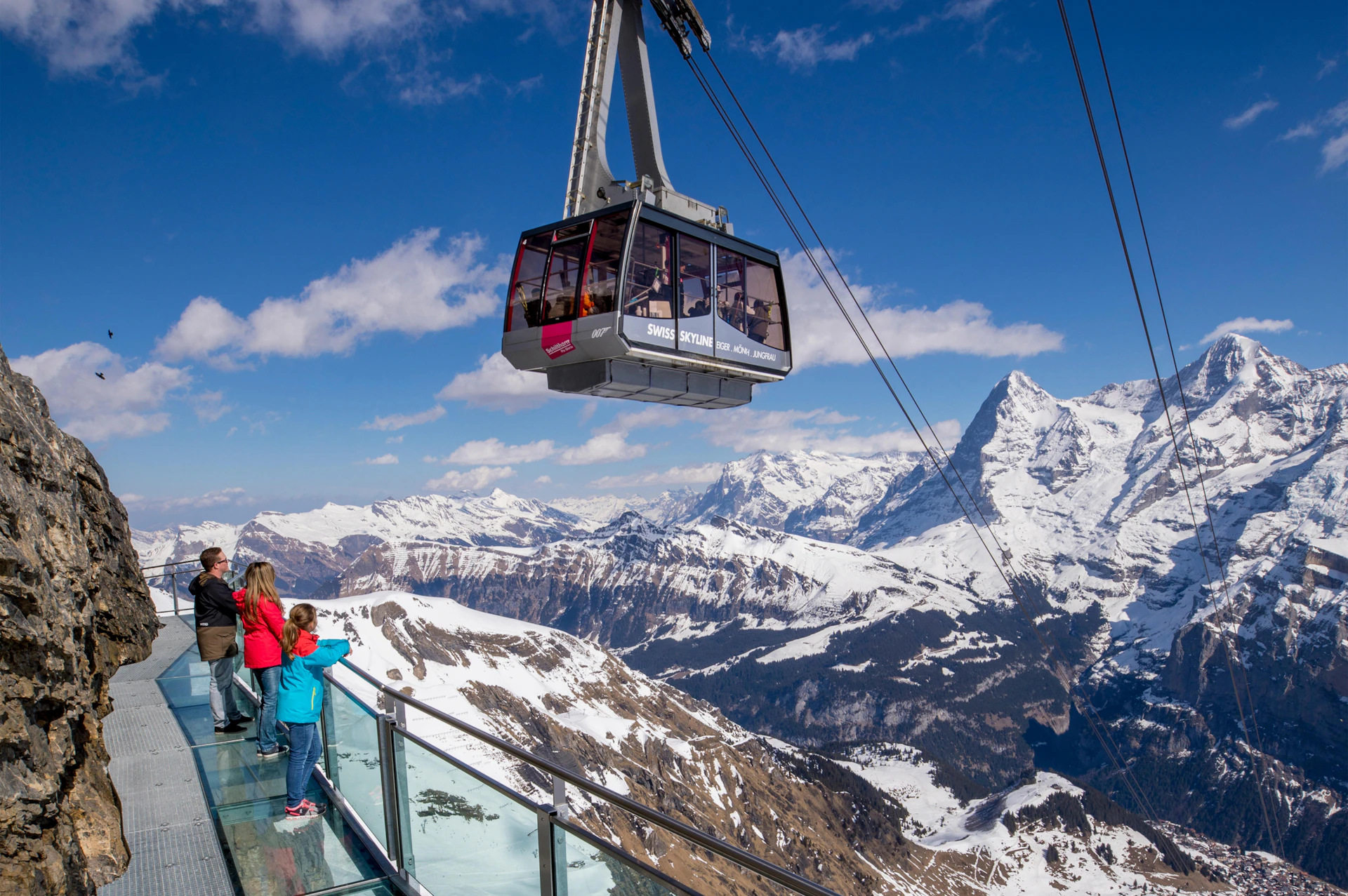 People stand on a glass platform overlooking snowy mountains as a cable car ascends under a clear blue sky.