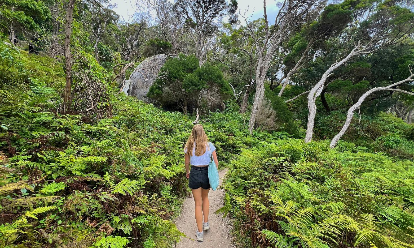 Μια γυναίκα περπατά σε ένα χωματόδρομο μέσα από πυκνή πράσινη βλάστηση σε μια δασώδη περιοχή με ψηλά δέντρα και θάμνους.