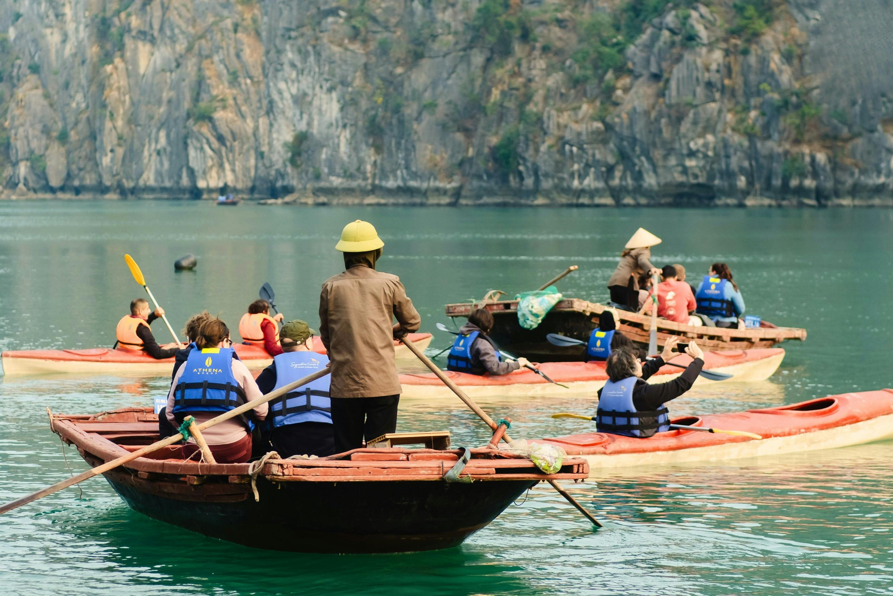 People wearing life jackets and paddling in wooden boats on a calm lake with a rocky shore in the background.