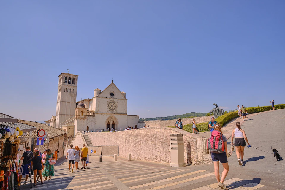 Tourists walking near the Basilica of Saint Francis in Assisi, Italy, under a clear blue sky.