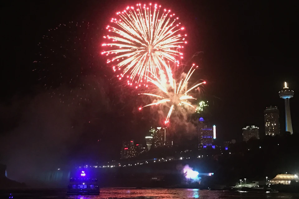 Bright fireworks burst over a city skyline at night with a boat illuminated by blue lights on the water below.