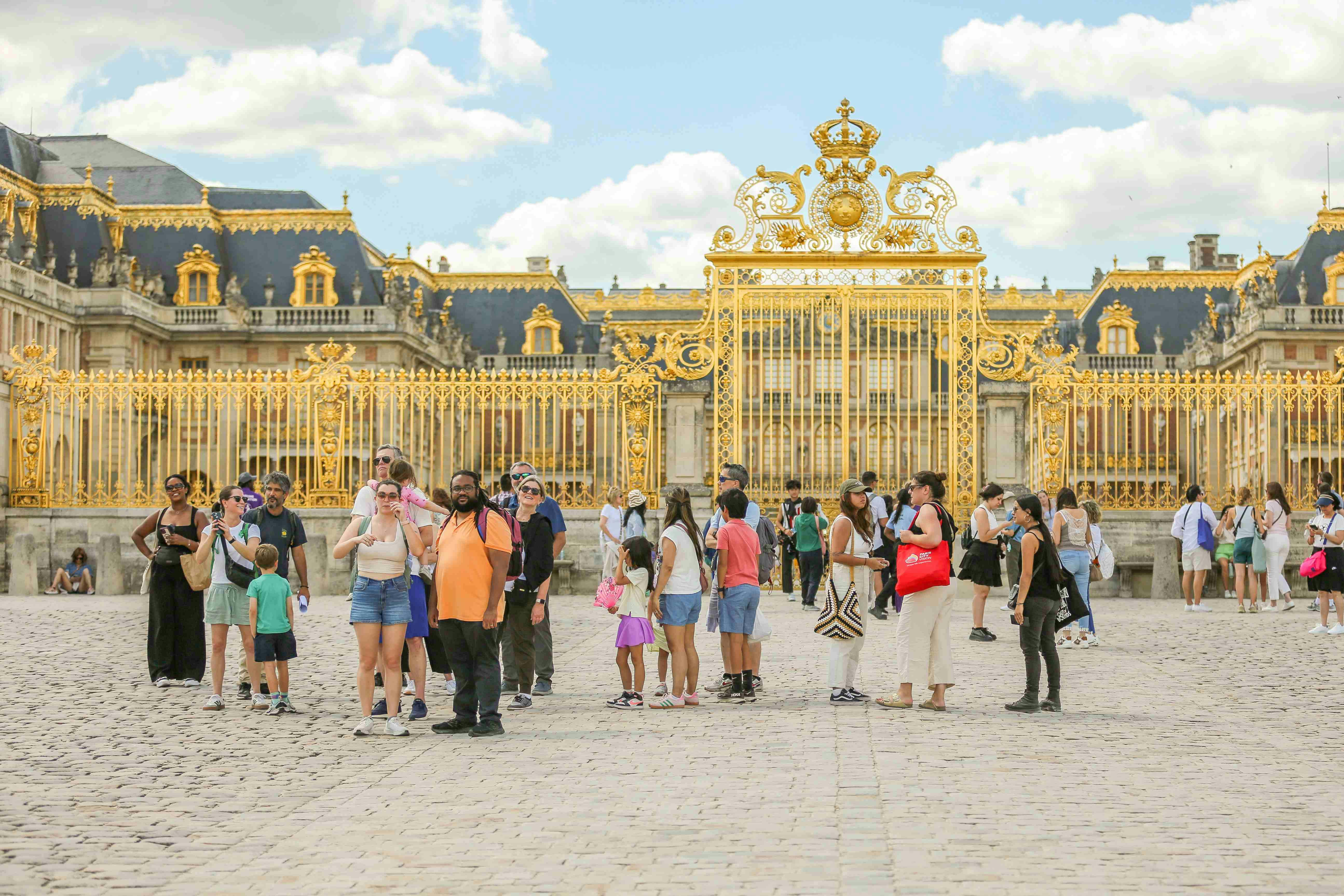 A group of tourists stands in front of ornate golden gates with a historic palace in the background under a partly cloudy sky.