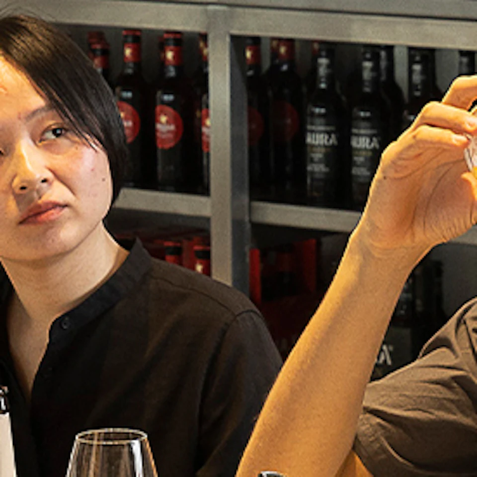 People seated at a table in a brightly lit room, tasting beverages, while a woman stands and talks. Shelves with bottles are behind them.