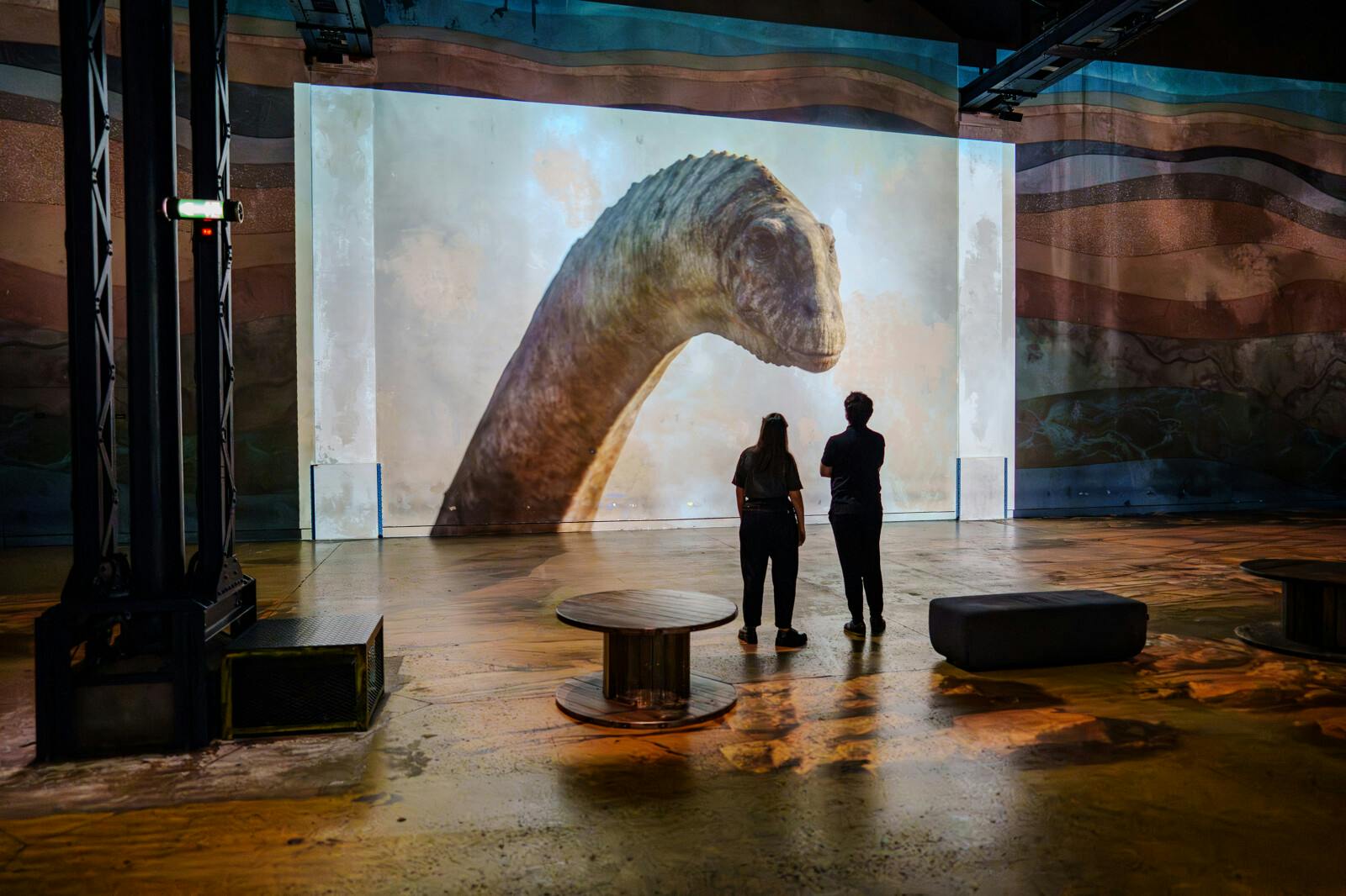 Δύο άνθρωποι στέκονται μπροστά από μια μεγάλη οθόνη που εμφανίζει την εικόνα του κεφαλιού και του λαιμού ενός δεινοσαύρου σε ένα σκοτεινό, ευρύχωρο δωμάτιο.