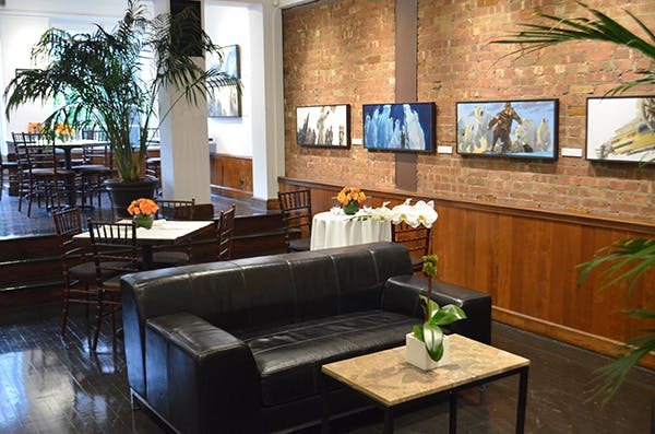 Contemporary lounge with black leather sofa, round and square tables, brick wall with paintings, and potted flowers.