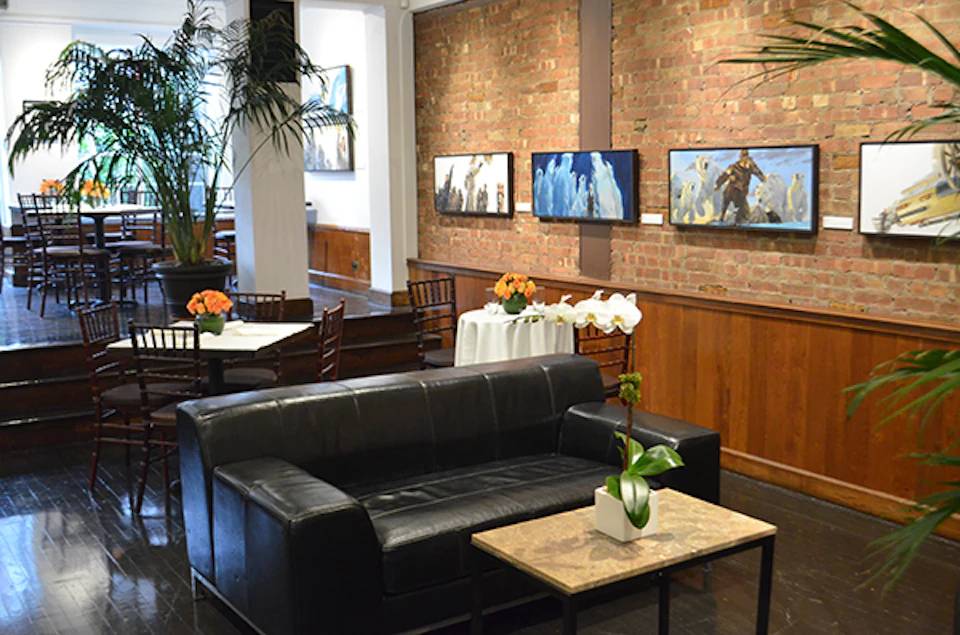 Contemporary lounge with black leather sofa, round and square tables, brick wall with paintings, and potted flowers.