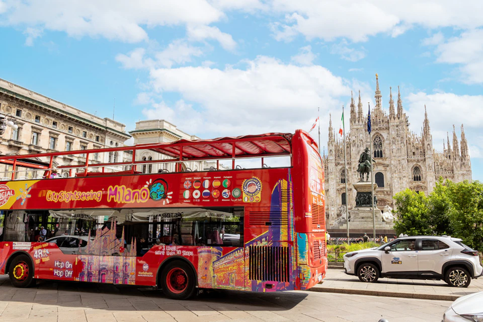 Red double-decker tour bus in Milan featuring colorful artwork, with the Milan Cathedral and a statue in the background under a blue sky.