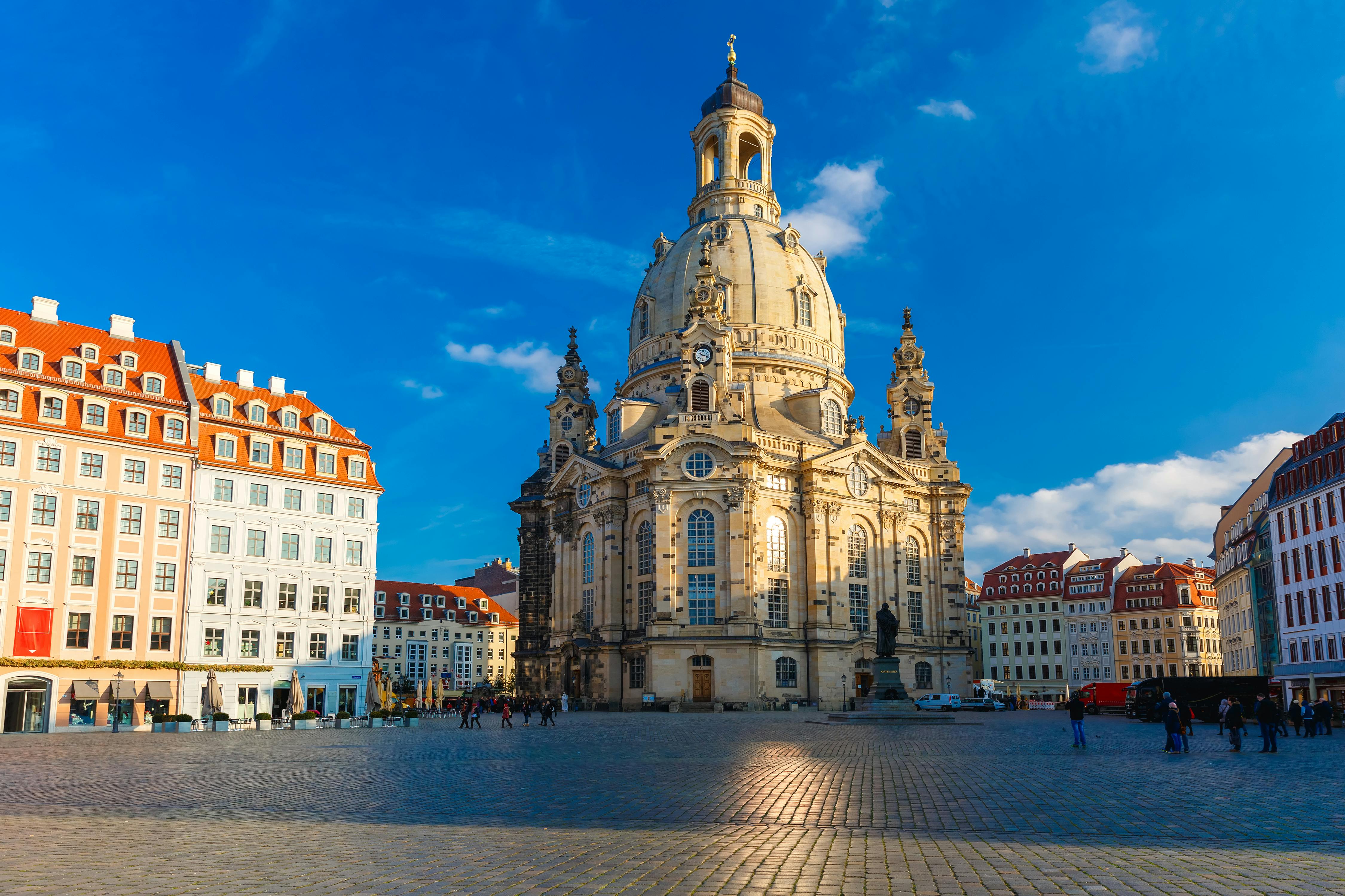 A large baroque-style church with a central dome stands in a sunlit square, surrounded by smaller buildings and a few people.