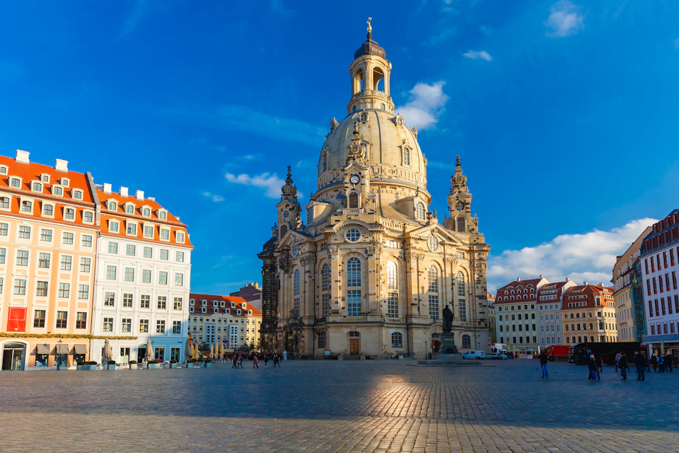 A large baroque-style church with a central dome stands in a sunlit square, surrounded by smaller buildings and a few people.