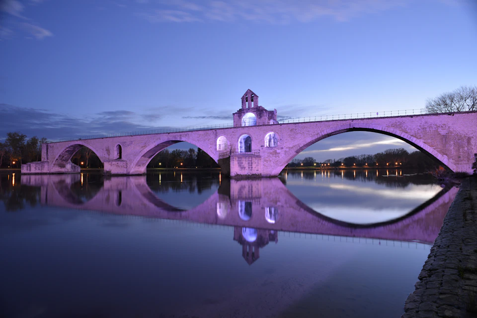Stone bridge with arches and a tower, illuminated in purple, reflecting in a calm river at dusk.