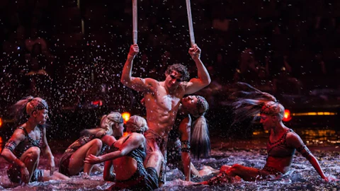 Performers in intricate costumes engage in a dynamic, water-themed act under dramatic lighting on a dark stage.