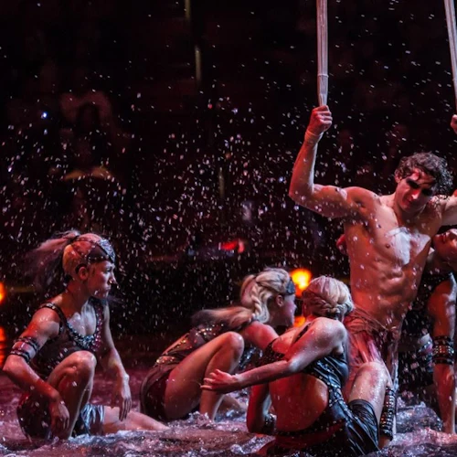 Performers in intricate costumes engage in a dynamic, water-themed act under dramatic lighting on a dark stage.