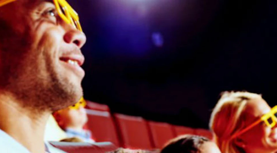 A group of people, including children, are wearing yellow 3D glasses and sitting in red theater seats, watching a screen.