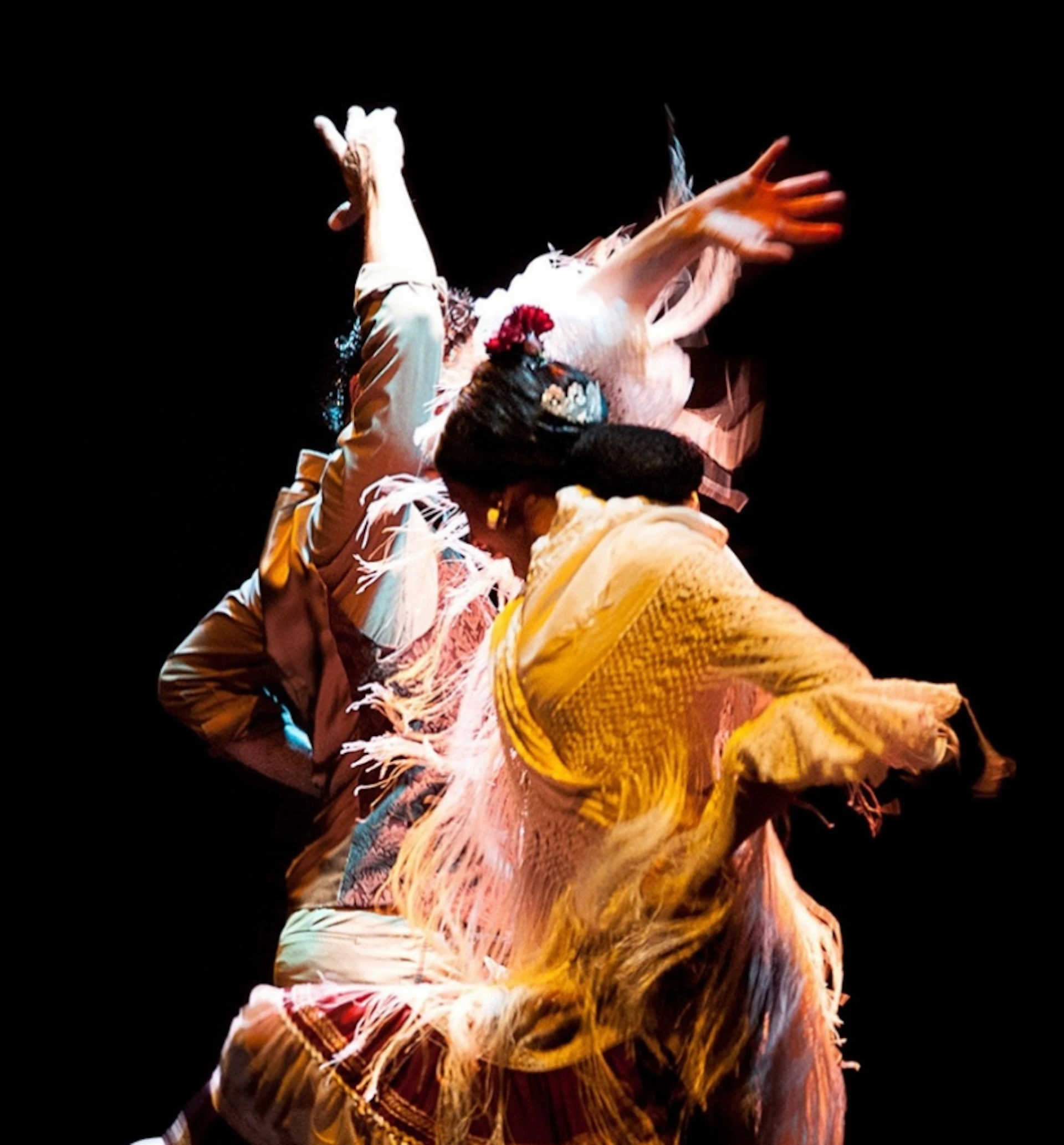 Two dancers in traditional attire perform a lively dance under dramatic lighting against a dark background.