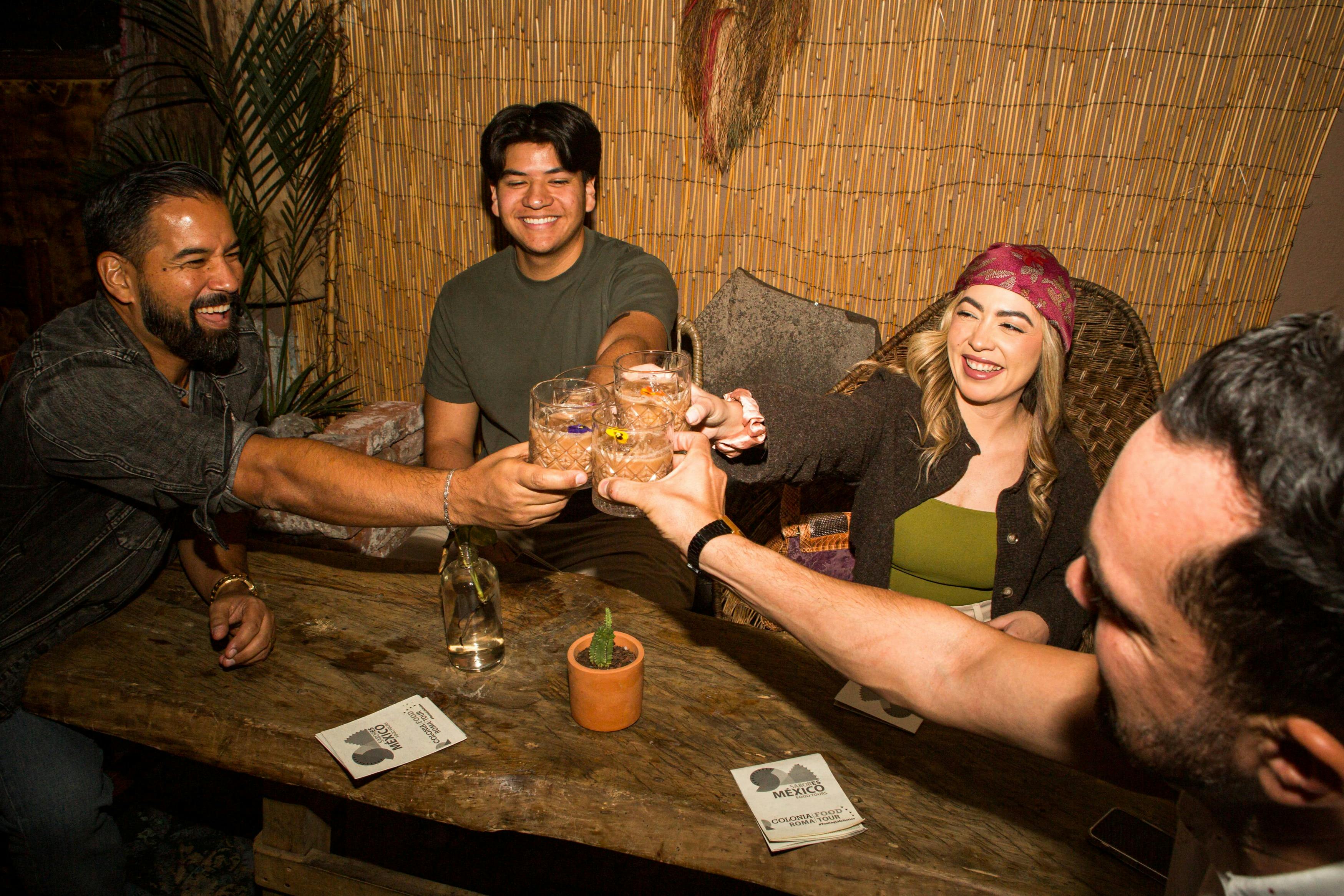 Un gruppo di quattro persone che sorridono e sorseggiano bicchieri insieme a un tavolo di legno con un piccolo cactus in vaso e sottobicchieri.