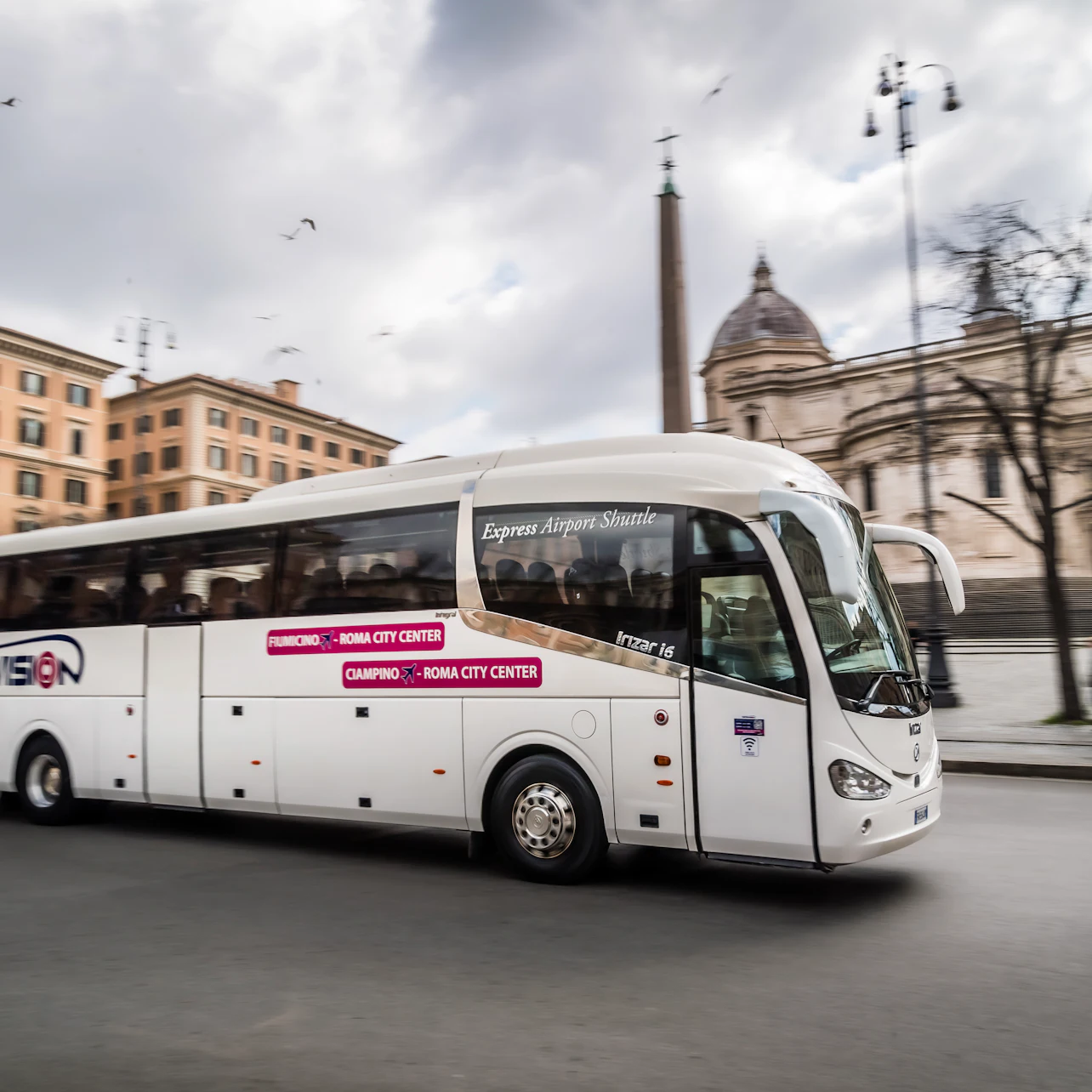 A white shuttle bus labeled "Express Airport Shuttle" driving in a city with historical buildings in the background.