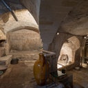 An ancient, stone-walled underground room with arched ceilings, a large ceramic pot, and minimal lighting.