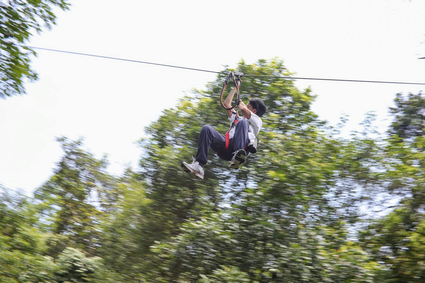 A person ziplining through a forested area, holding onto the harness, with trees blurred in the background.