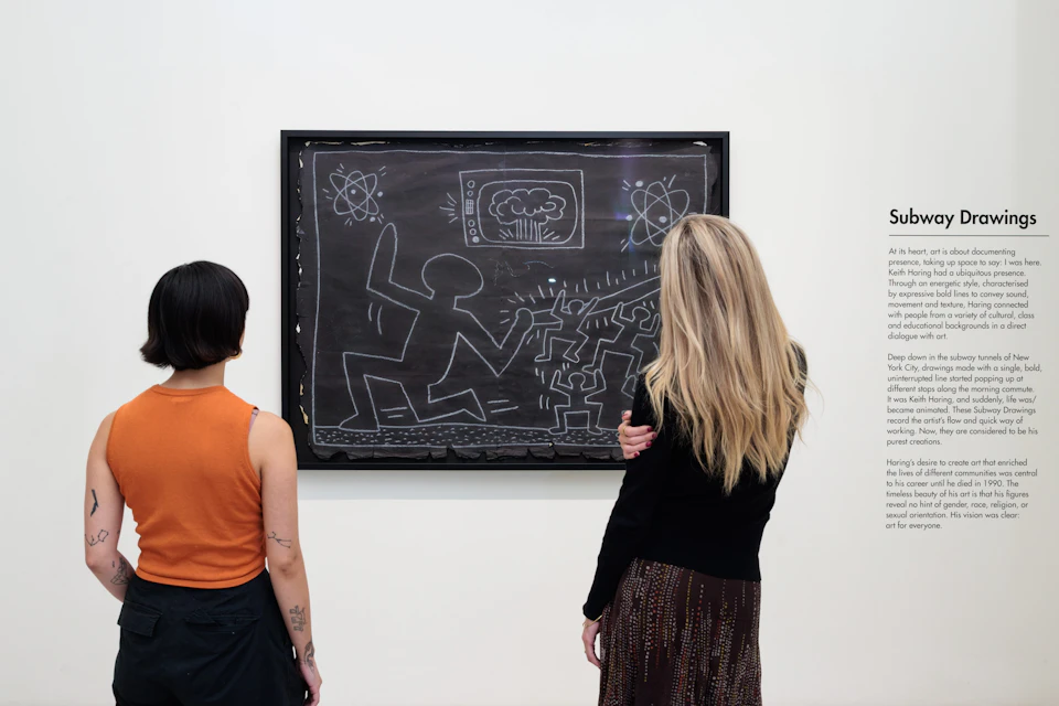 Two people observe a framed artwork on a wall, featuring chalk-like drawings of figures, shapes, and symbols on a black background.