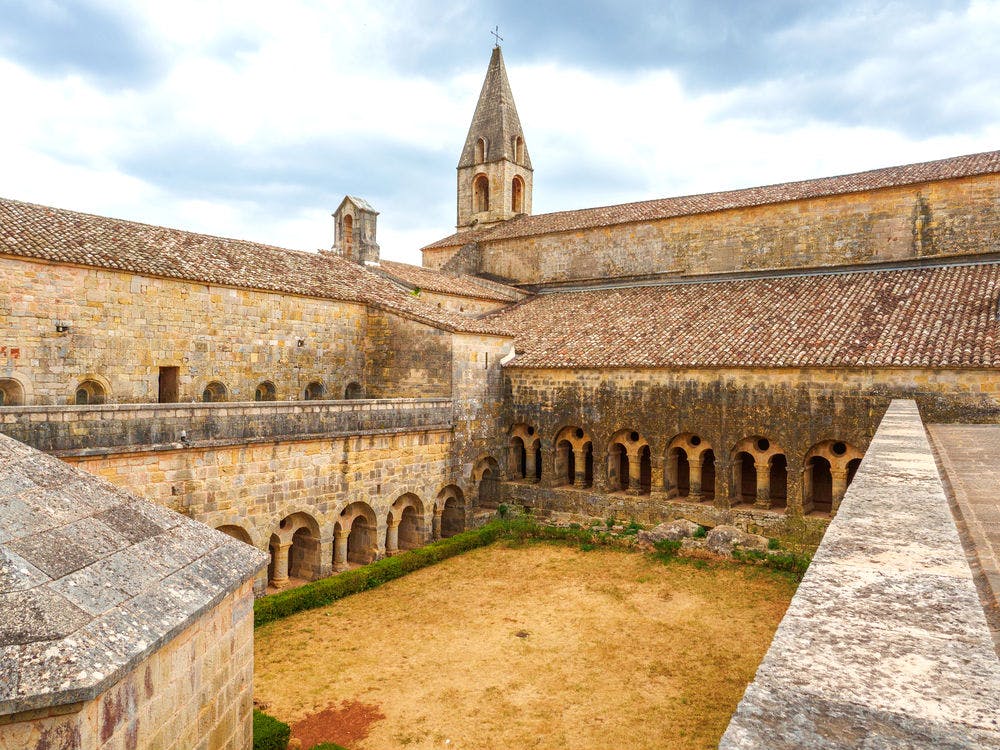 Αρχαία πέτρινη αυλή μοναστηριού με τοξωτούς πεζόδρομους, μια κεντρική ξερή χλοώδη περιοχή και ένα ψηλό κωδωνοστάσιο κάτω από ένα συννεφιασμένο ουρανό.