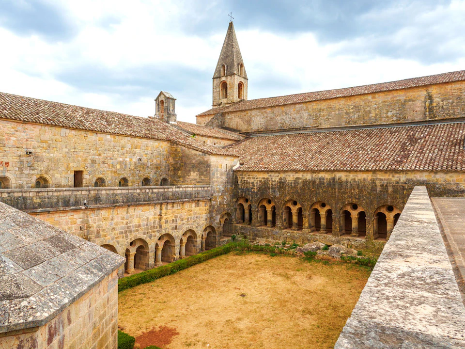Ancient stone monastery courtyard with arched walkways, a central dry grassy area, and a tall spire under a cloudy sky.