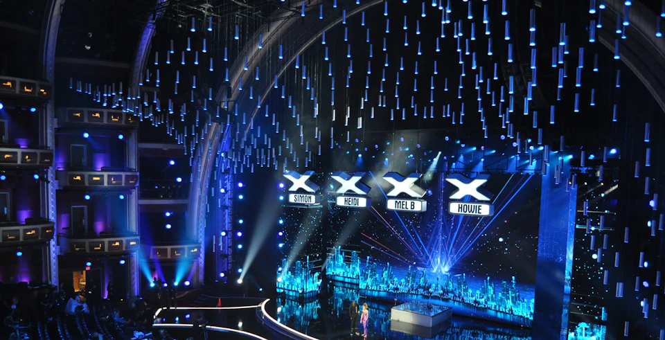 Stage with blue lighting, large X symbols above labeled "Simon," "Heidi," "Mel B," and "Howie." Two hosts stand below them.
