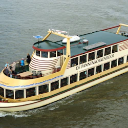 En båt som heter "De Pannenkoekenboot" på vatten med människor på däck och en holländsk flagga baktill.