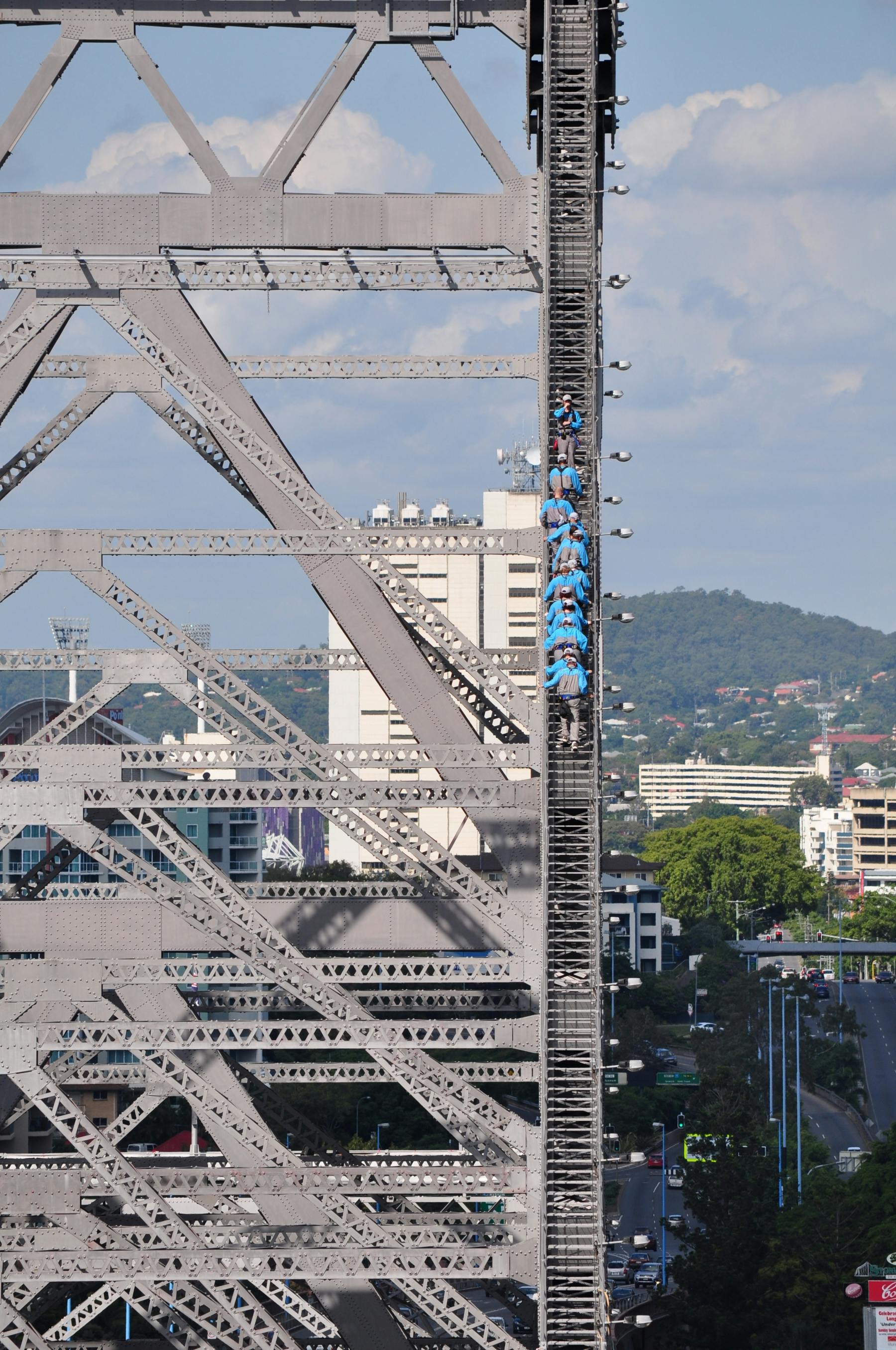 一群身穿蓝色衬衫的人攀登着一座金属桥梁结构，背景是城市景观和山丘。